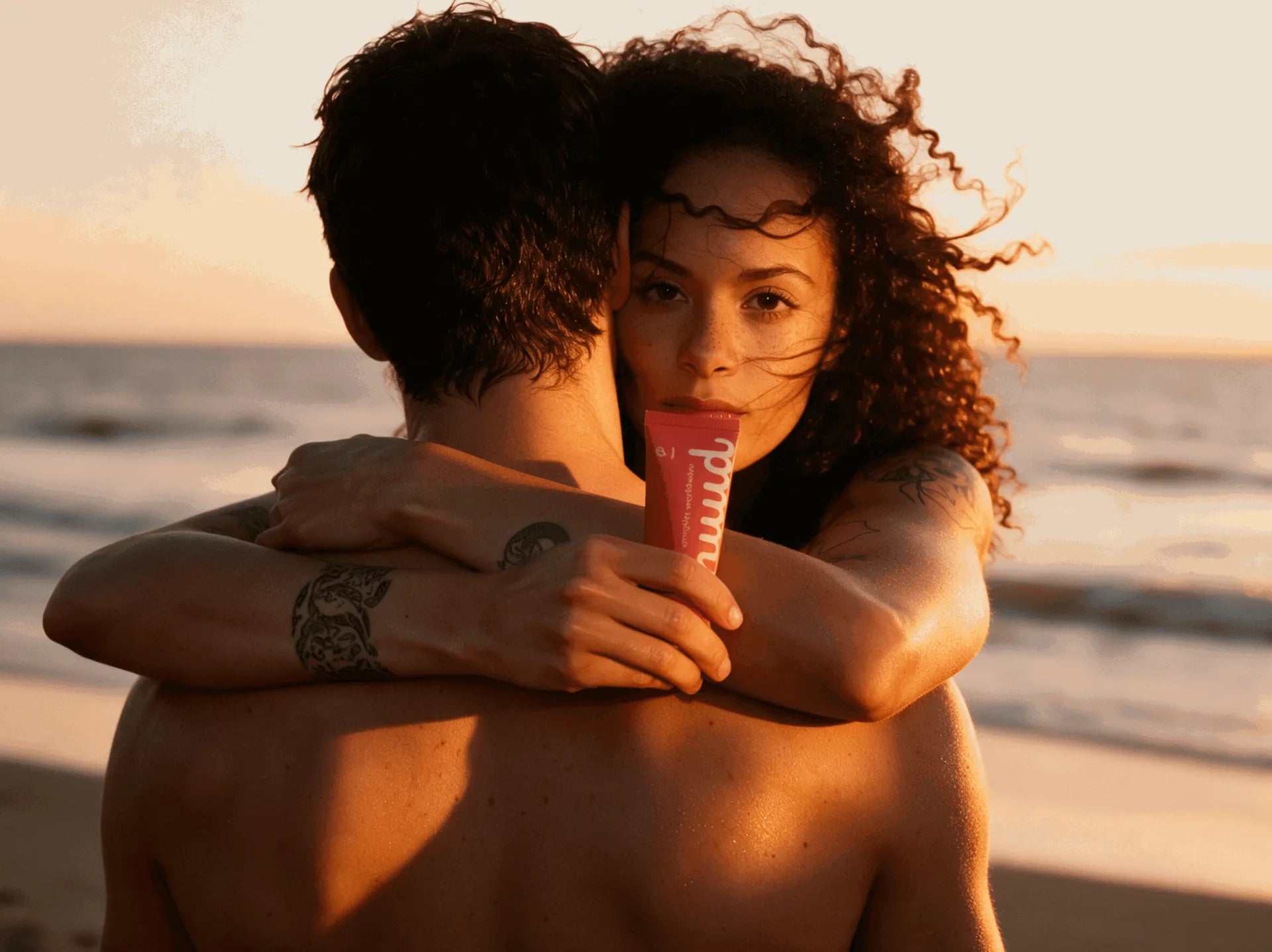 A woman embracing someone on a beach at golden hour, holding a tube of nuud